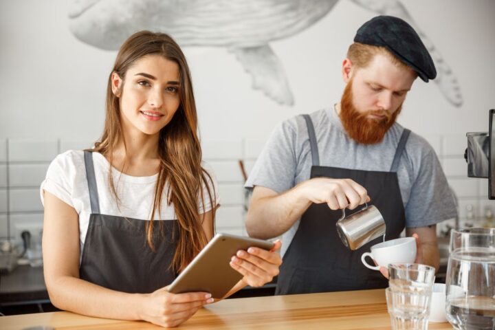 Two baristas are behind the counter in a café, one is holding a tablet looking directly to camera whilst the other pours coffee