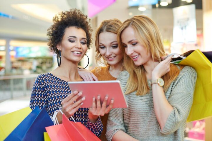 Three friends in a mall gather round a tablet whilst holding lots of shopping bags